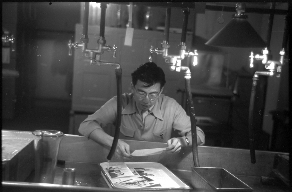 Photographic negative of Frank Hirahara sitting in front of a large sink developing photographs. Courtesy of Frank C. Hirahara Collection, Japanese American Museum of Oregon; Densho.