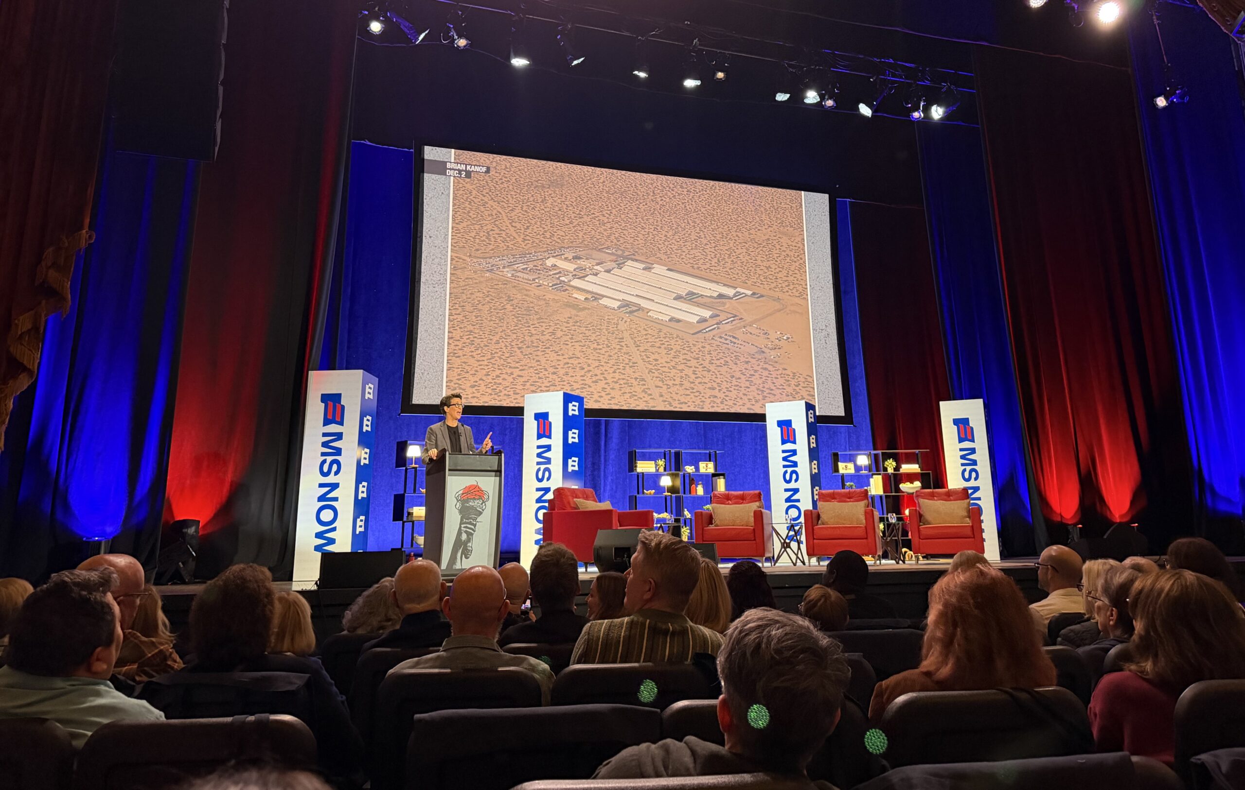 Rachel Maddow speaking on stage with image of detention camp projected on screen in background.
