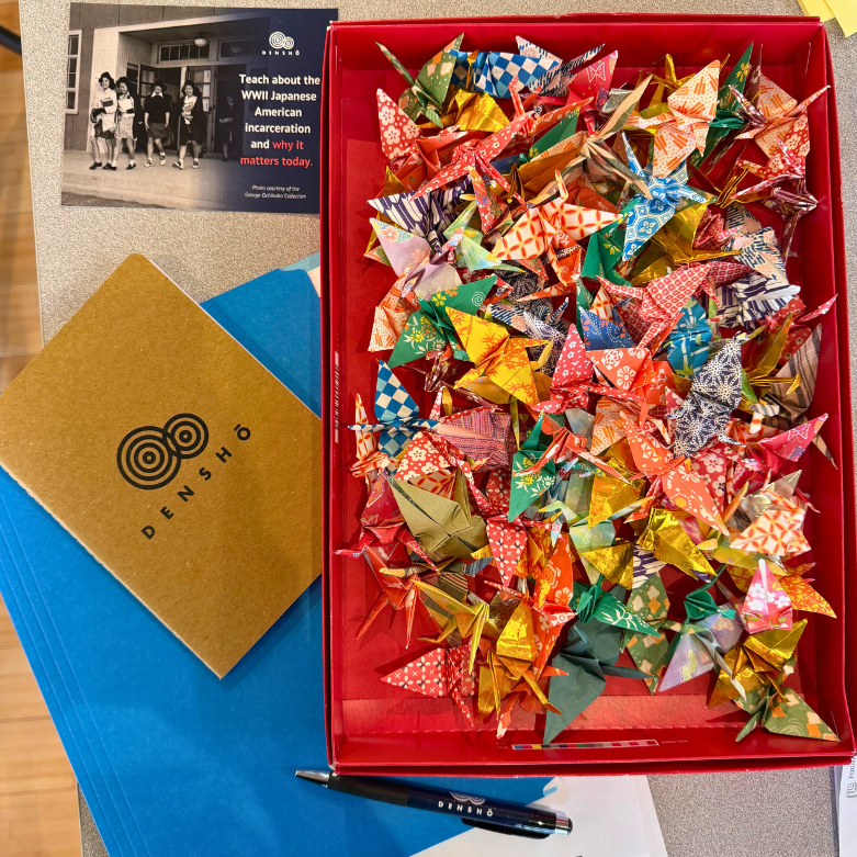 A blue folder, Densho notebook, an open box of multicolored, origami cranes, and a descriptive postcard about Densho
