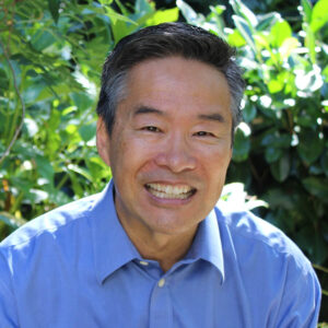 Tom Ikeda headshot; Tom standing in front of green foliage background