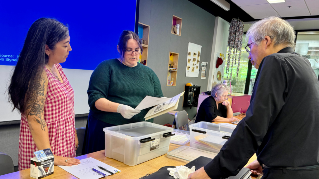 Two Densho archivists stand on the left hand side across from a community member standing opposite them on the other side of a desk. The Densho staff hold and review papers, talking about them with the community member.