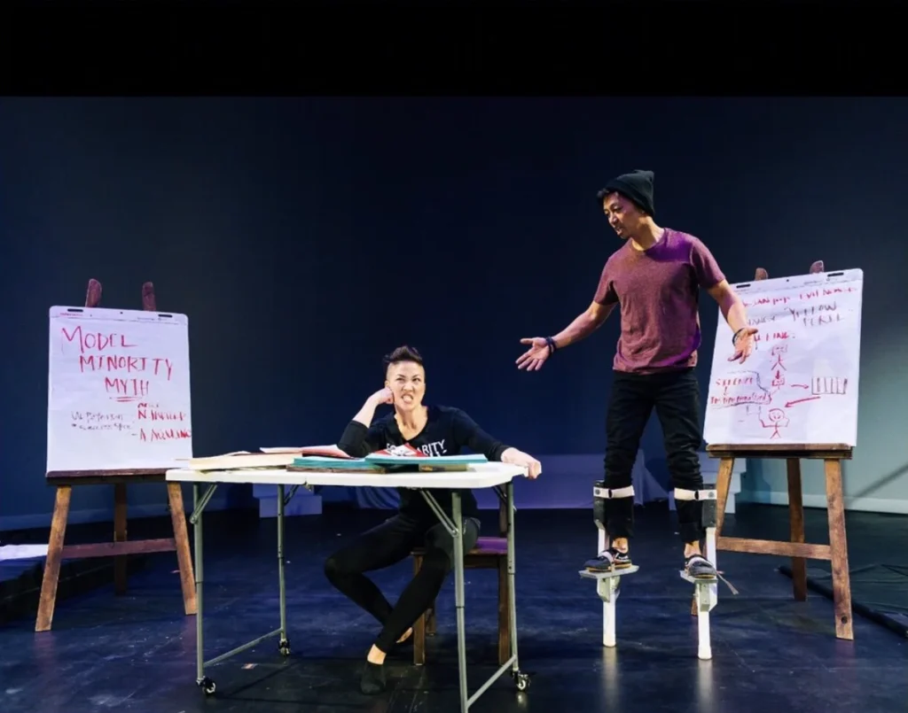 Photo of two actors on stafe -- one Asian American woman on the left sitting in a thinking position at a desk, looking frustrated, with an Asian American man to standing to her right, who is talking enthusiastically. The actors are on stage, and there are two hand-written posters propped up next to them. One poster says "Model Minority Myth" and the other says "Yellow Peril" and has a stick-figure drawing.