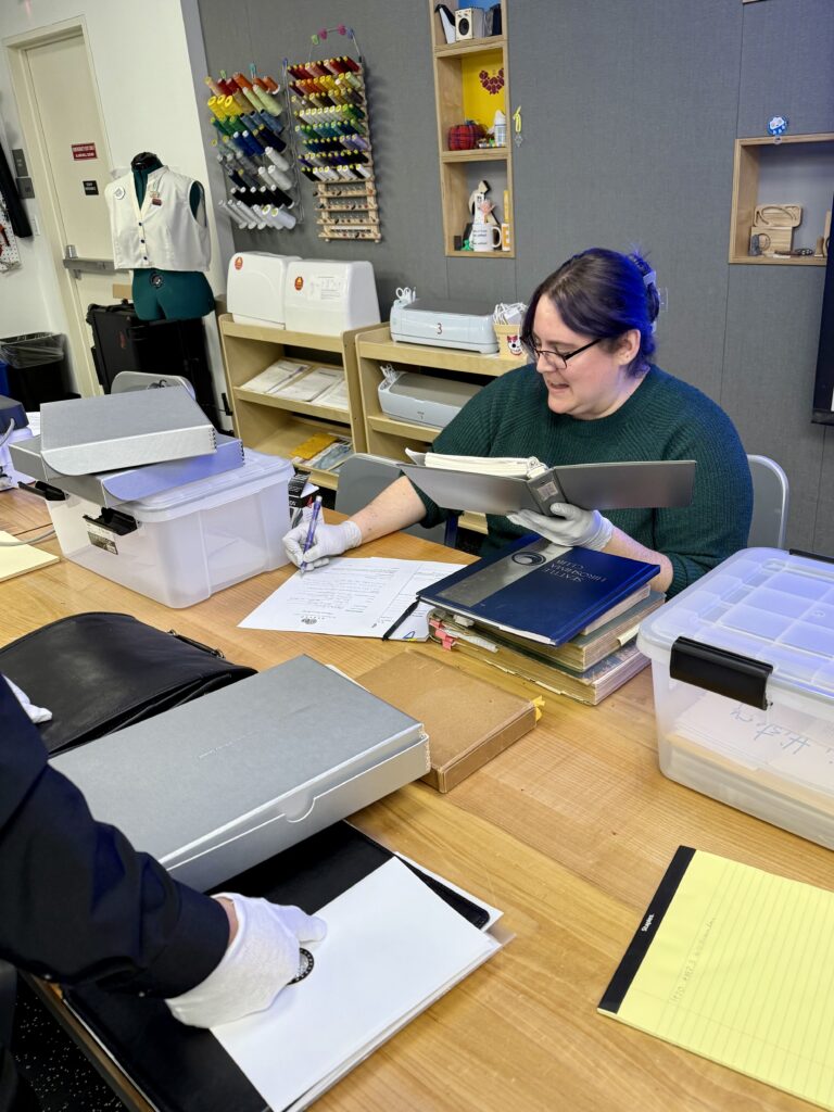 Densho archivist Micah Merryman sits at a desk and looks through a donor’s collection. She is holding a book in one hand and taking notes on a paper with the other hand. On the desk sits a stack of books, as well as a silver box, a small brown box, and some other materials.