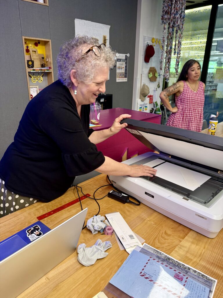 Densho archivist Dina Moreno is standing and using a flatbed scanner, which is placed on a wooden desk. Next to the scanner are papers and her laptop.