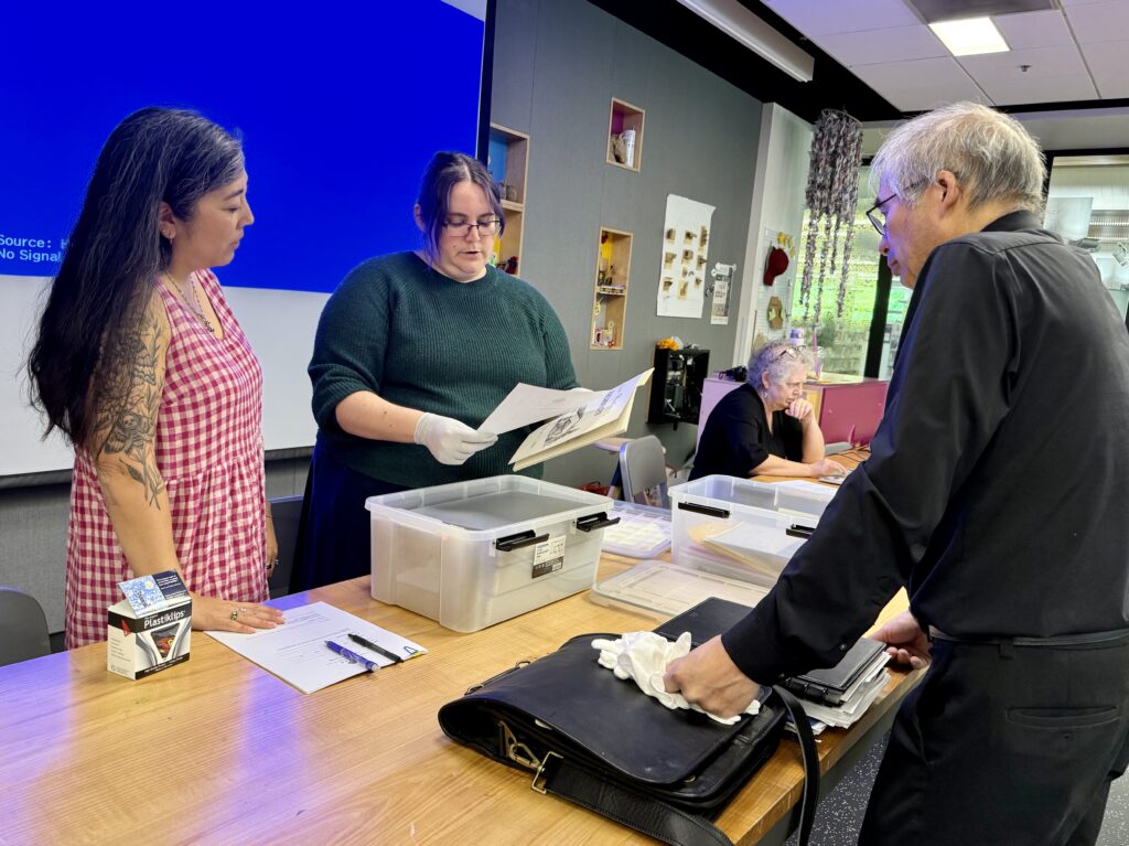 Two Densho archivists stand on the left hand side across from a community member standing opposite them on the other side of a desk. The Densho staff hold and review papers, talking about them with the community member.