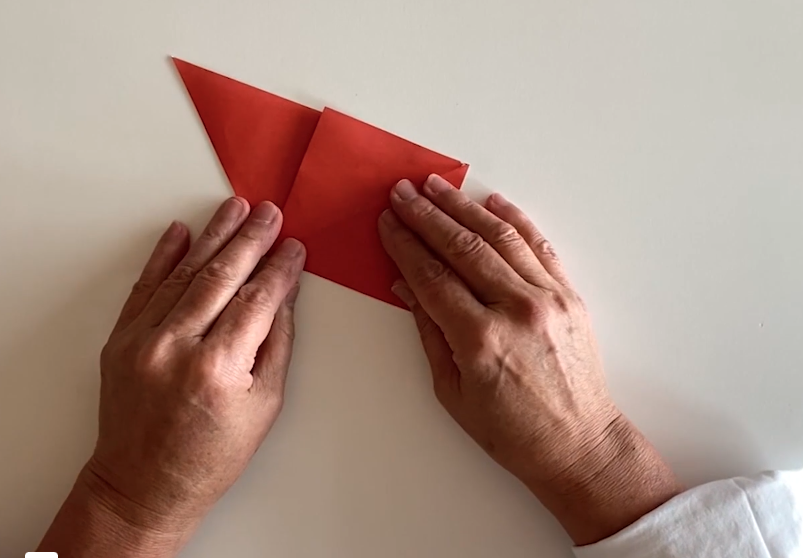 A photo of two hands (likely of a middle aged person) folding a piece of red origami paper.