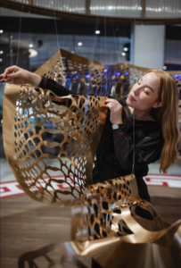 Photo of woman holding piece of paper cutting artwork. The paper art is golden and cut into a shape like fish scales, and it stretches into the background.