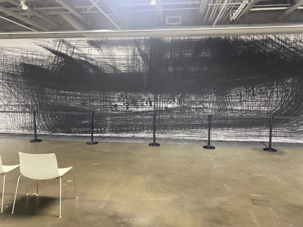 A photo of an art exhibit. A large white canvas is stretched across a wall, and black painted lines are layered hundreds of times on top of the white background. The lines vaguely resemble the shape of a ship. In front are two empty chairs for viewing the artwork.