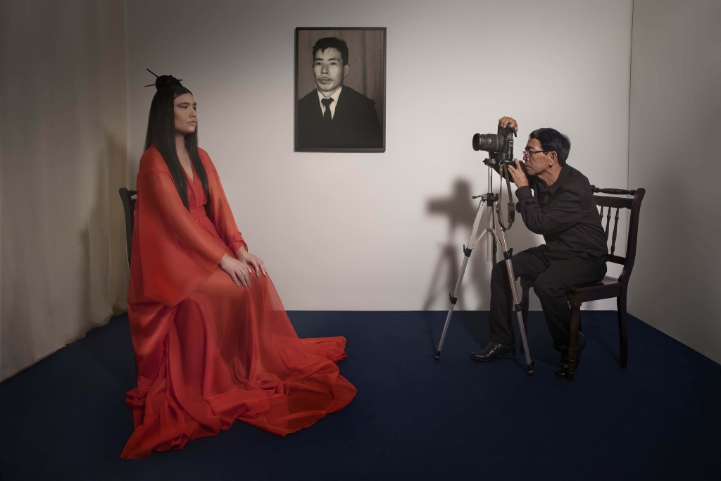 A photo of an Asian man dressed in black on the right hand side taking a photo of a woman wearing a red silk kimono on the left. A portrait of an Asian man hangs on the wall in the background between them.