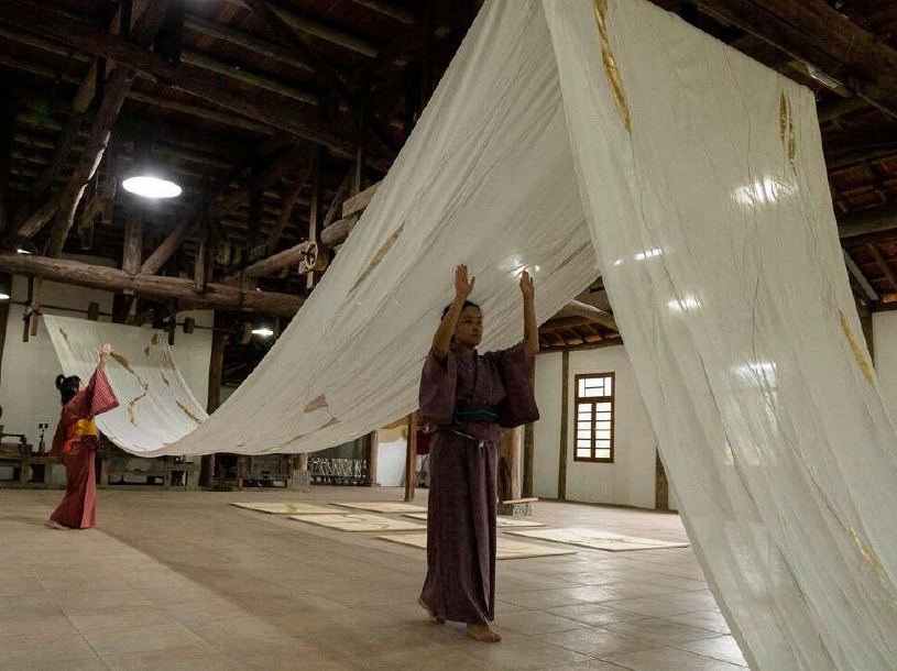 Photo of artwork. One woman in the foreground is wearing a Kimono, and she walks with her hands up underneath a large piece of white fabric with gold highlights hanging over her. A second woman in the background is also wearing a kimono and walking toward the large white hanging fabric, which stretches from the back wall and forward out of the picture frame.