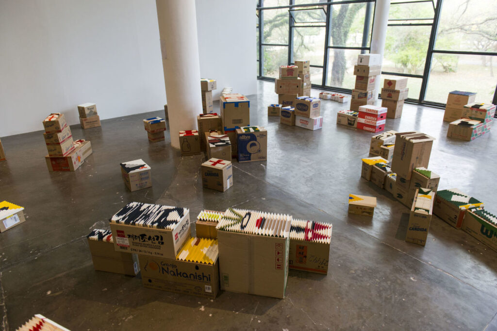 Photo of an art exhibit. Various cardboard boxes are strewn across a wide open room, with a big window in the background. Some boxes are stacked, others are alone, and they have artistic folded paper on top of them.