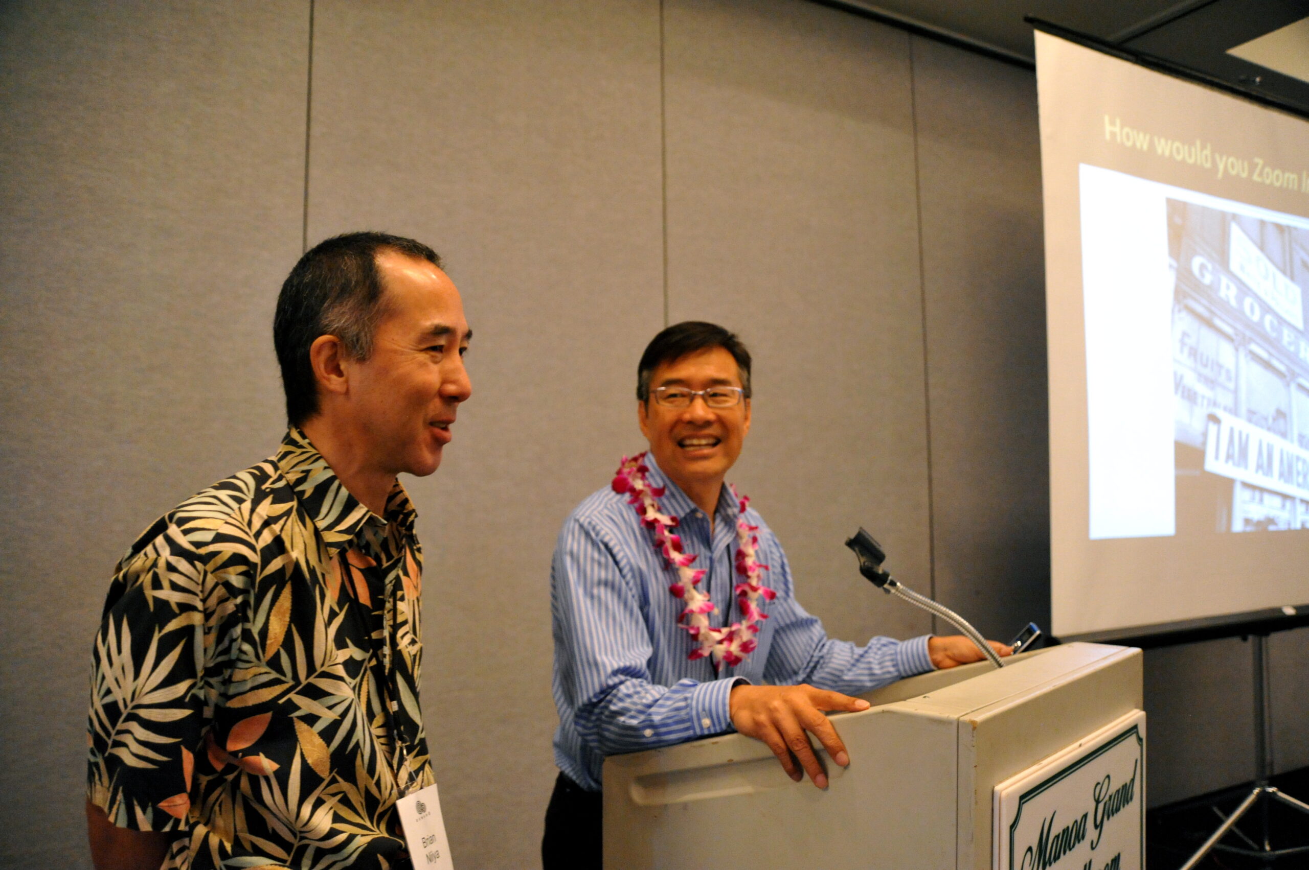 Two smiling men stand at the front of a room, with a projector off to one side. The project shows an image of a black and white archival photo featuring the words "I Am an American."