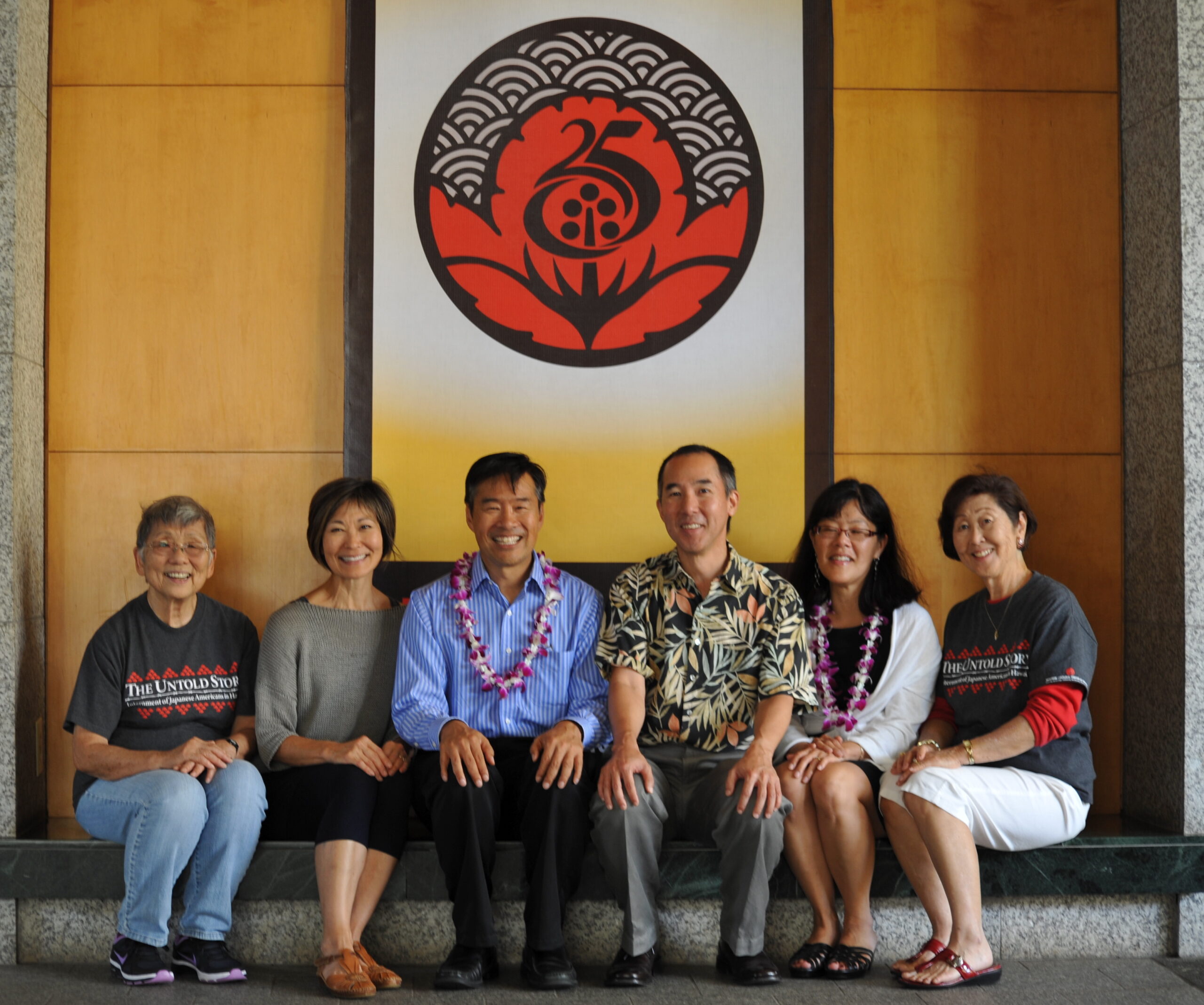 Group photo of six people seated in a lobby, smiling for the camera. Above them hangs a large black and red sign with with Japanese motifs and the number 25. Two people wear purple orchid leis.