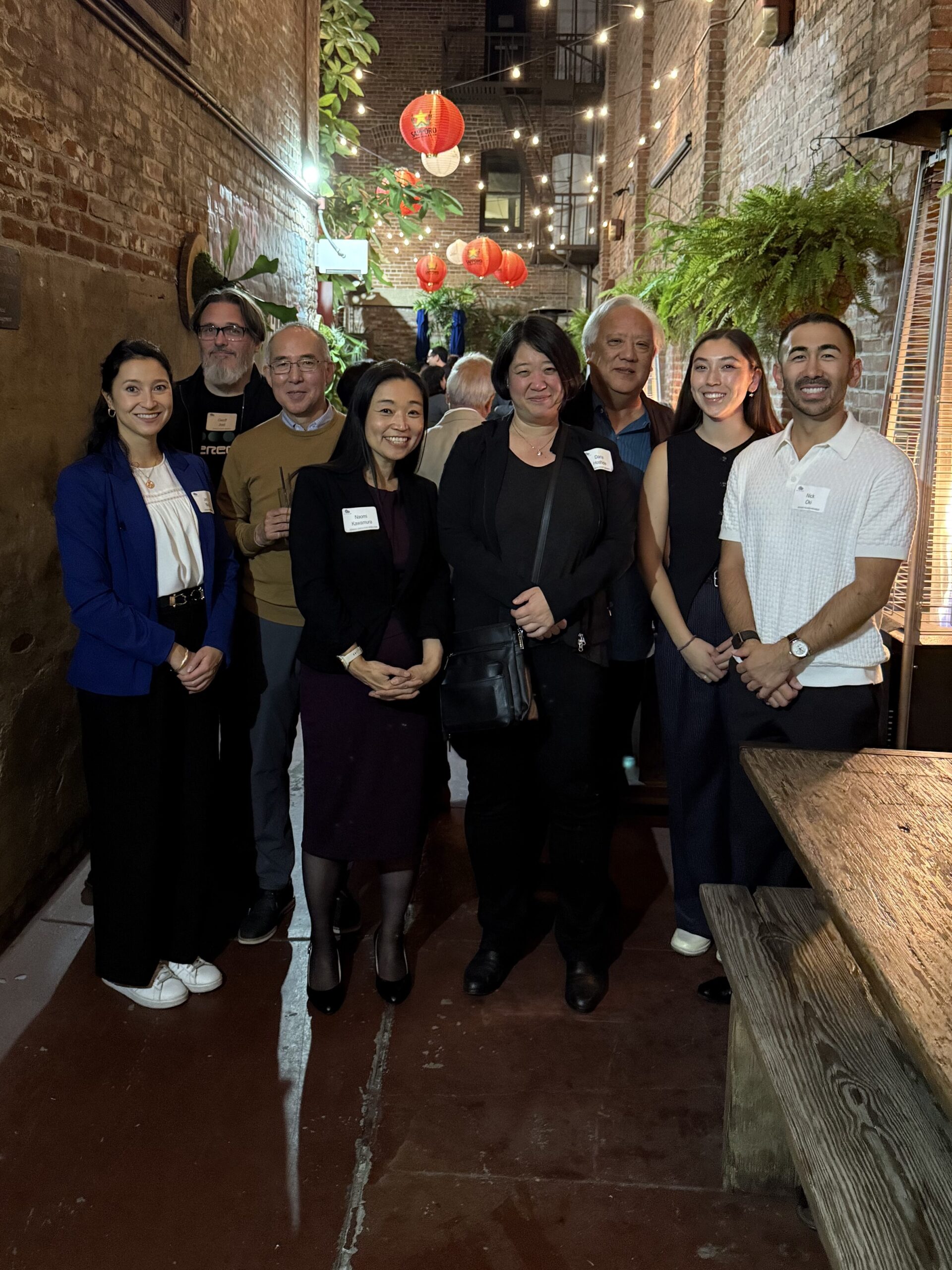 A group photo featuring eight people standing between brick buildings at night with Japanese chochin lanterns lit above them in the background. Everyone is smiling and facing the camera.