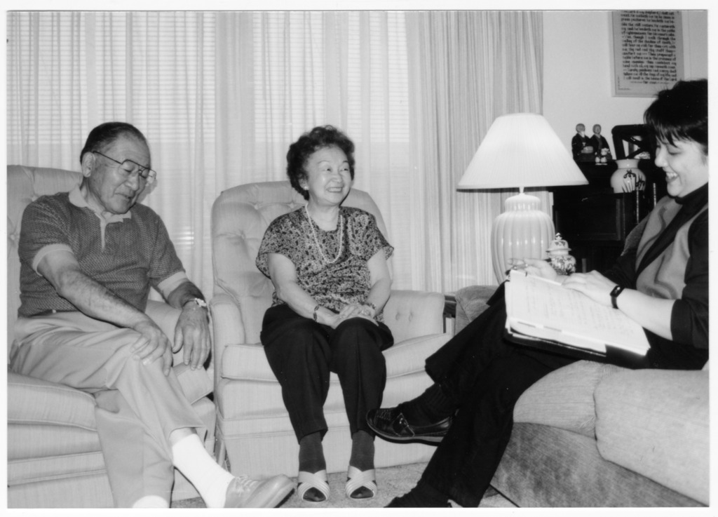 Black and white photo of elderly man and woman sitting on a couch, being interviewed by younger woman.