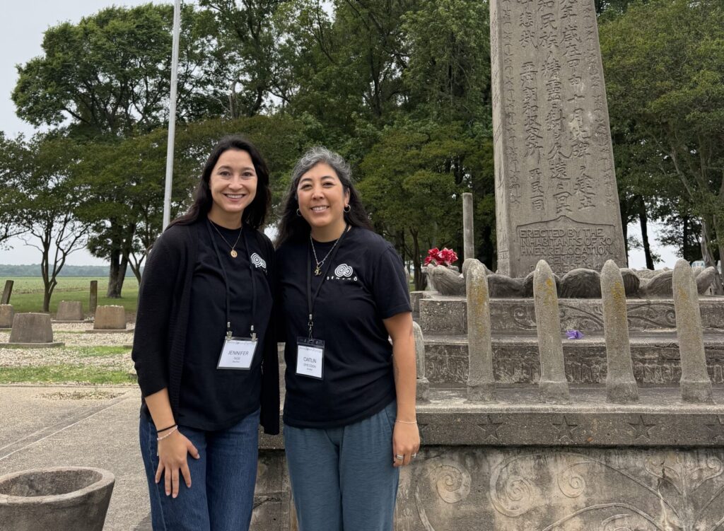 Two women stand smiling in front of a tall stone memorial engraved with Japanese text, with an American flag flying beside them.