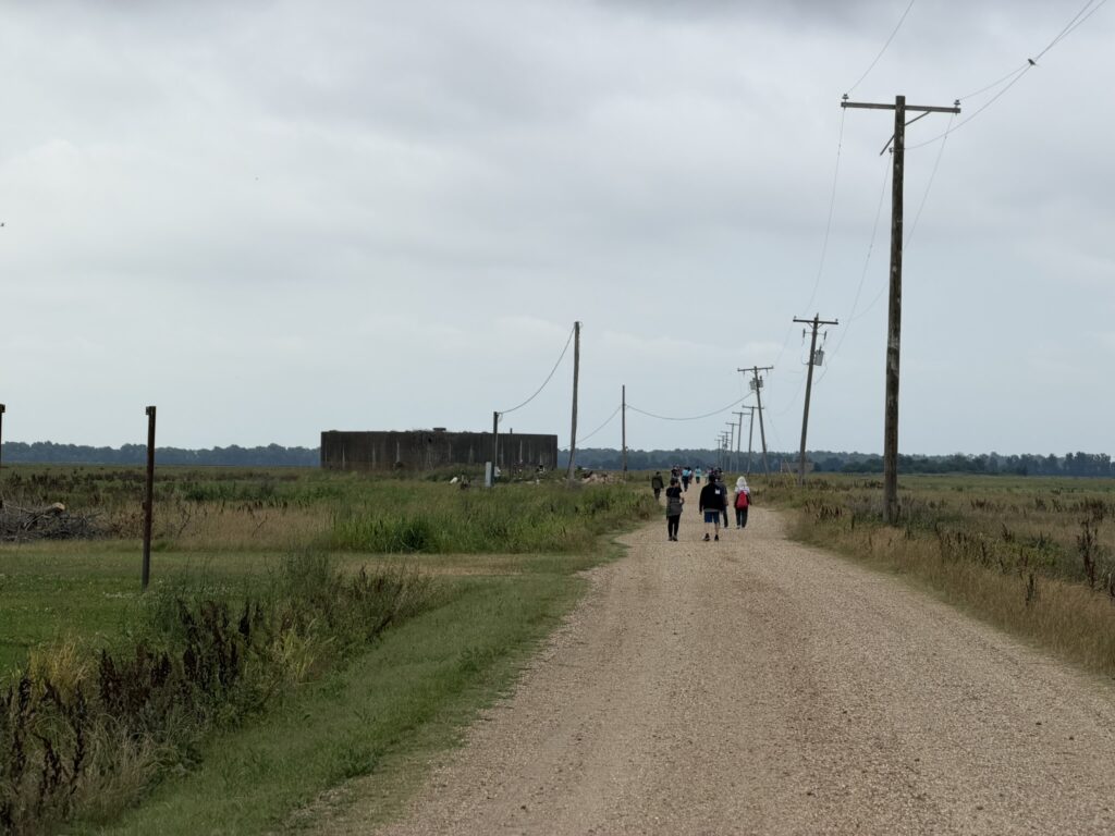 A group of people walks down a long gravel road lined with utility poles, passing a low concrete structure in an open field.