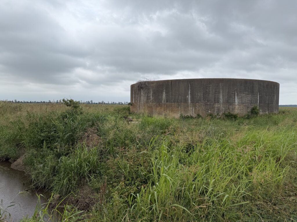 A large circular concrete water tank stands in an overgrown field bordered by tall grass and a small creek.