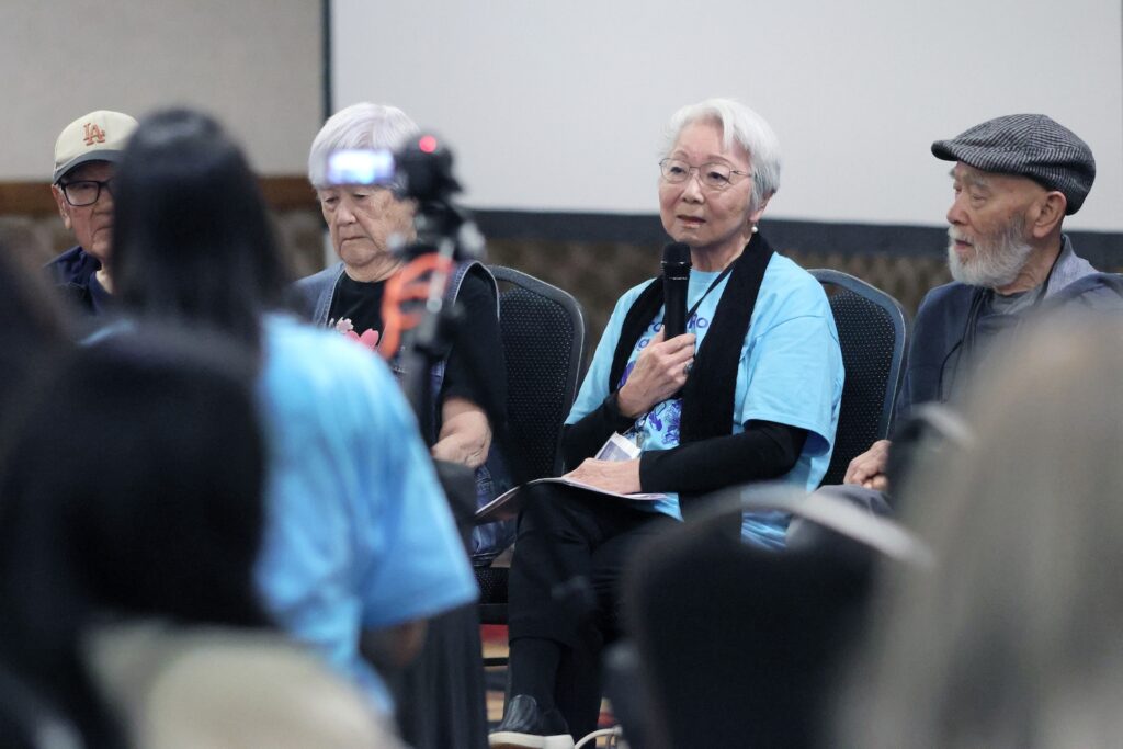 Four elderly seated panelists, two men and one women, participate in a discussion; a woman in the center speaks into a microphone.