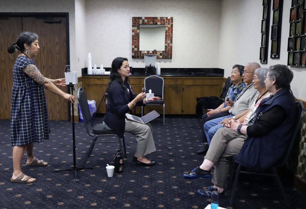 A woman holds a phone on a small tripod to record an interview. Woman interviewer sitting in front facing four people who are being interviewed in a hotel meeting room.