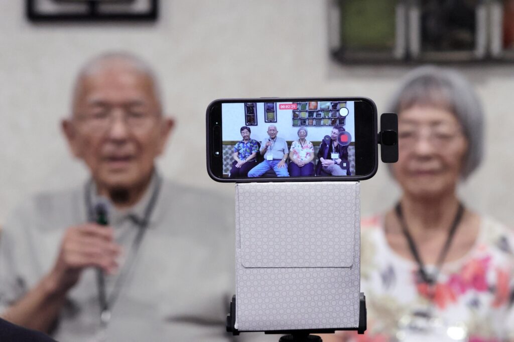 An out-of-focus older man and woman sit in the foreground while a phone screen mounted on a tripod displays a live video of four people being interviewed.