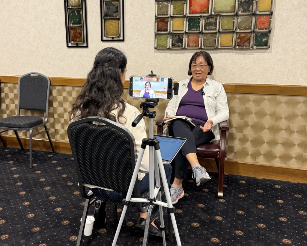 A woman interviewer sits facing away toward an older woman being interviewed in a hotel meeting room.