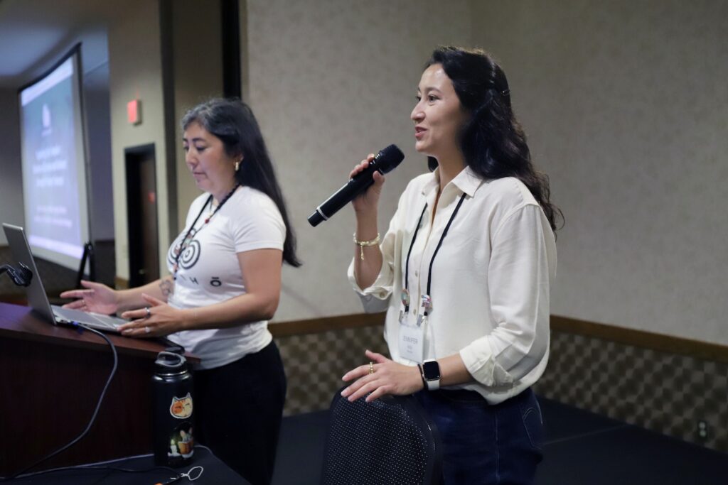 Two women stand at the front of a conference room giving a presentation; one holds a microphone while the other looks at a laptop, with a slide projected on the screen behind them.