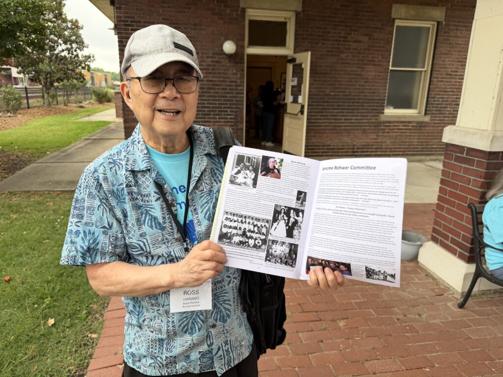 A smiling man wearing a name badge reading "Ross Harano" holds open a printed program showing photos and text about the Jerome Rohwer Committee, standing outside the museum.
