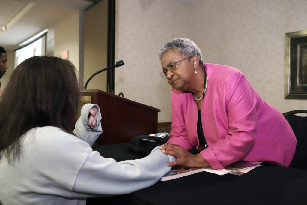 An older woman leans over a table to clasp hands warmly with a seated person, near a podium in a meeting room.