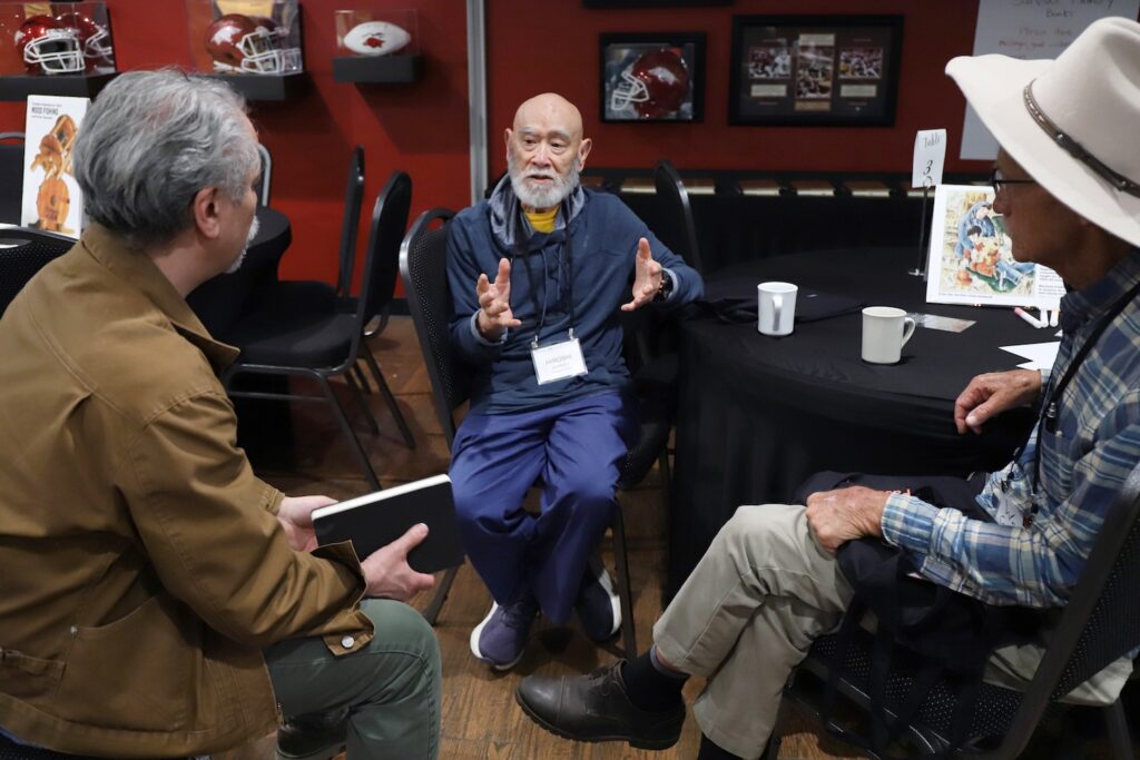 Three men sit and talk together at a small table in a room decorated with sports memorabilia, one gesturing with his hands as he speaks.