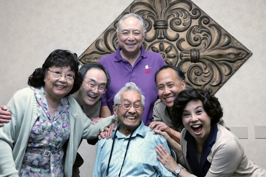 A group of six people, including an elderly seated man, smile and pose closely together indoors in front of a decorative wall panel.