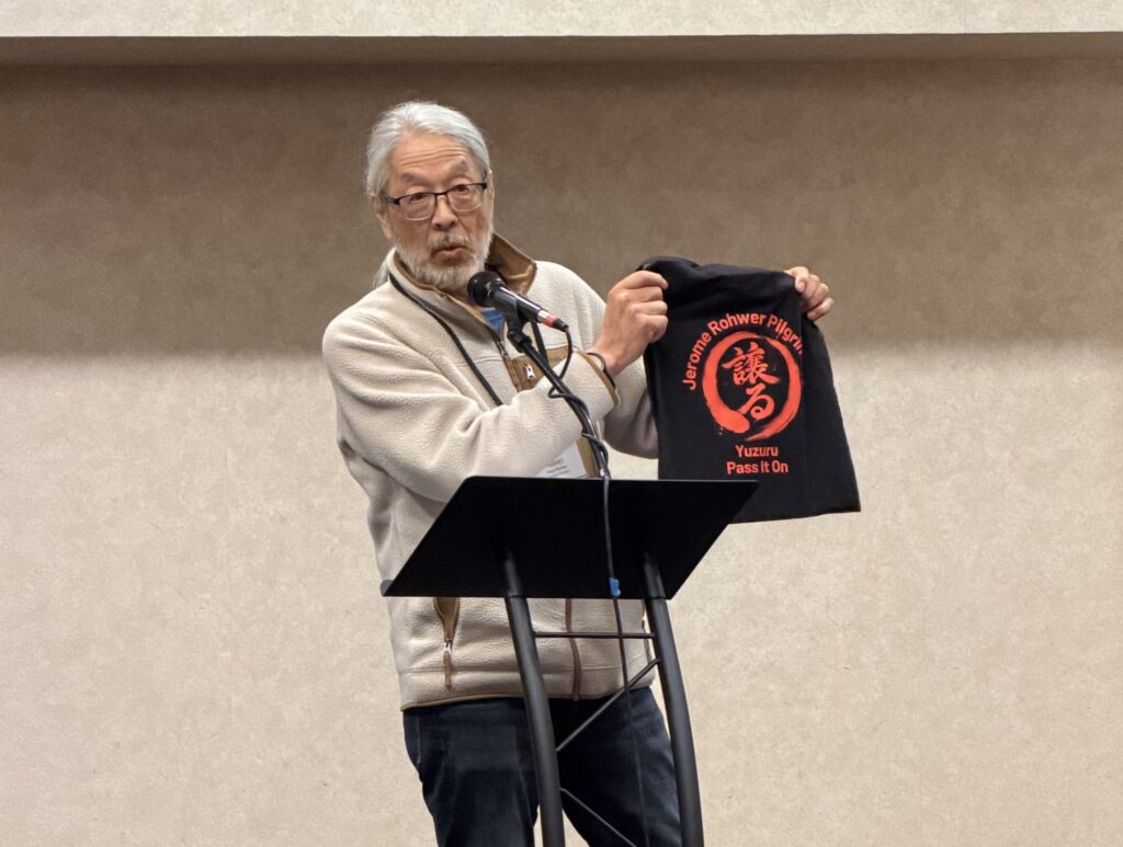 A man stands at a podium holding up a black tote bag printed with red Japanese characters for "Yuzuru" and the English words "Pass It On."