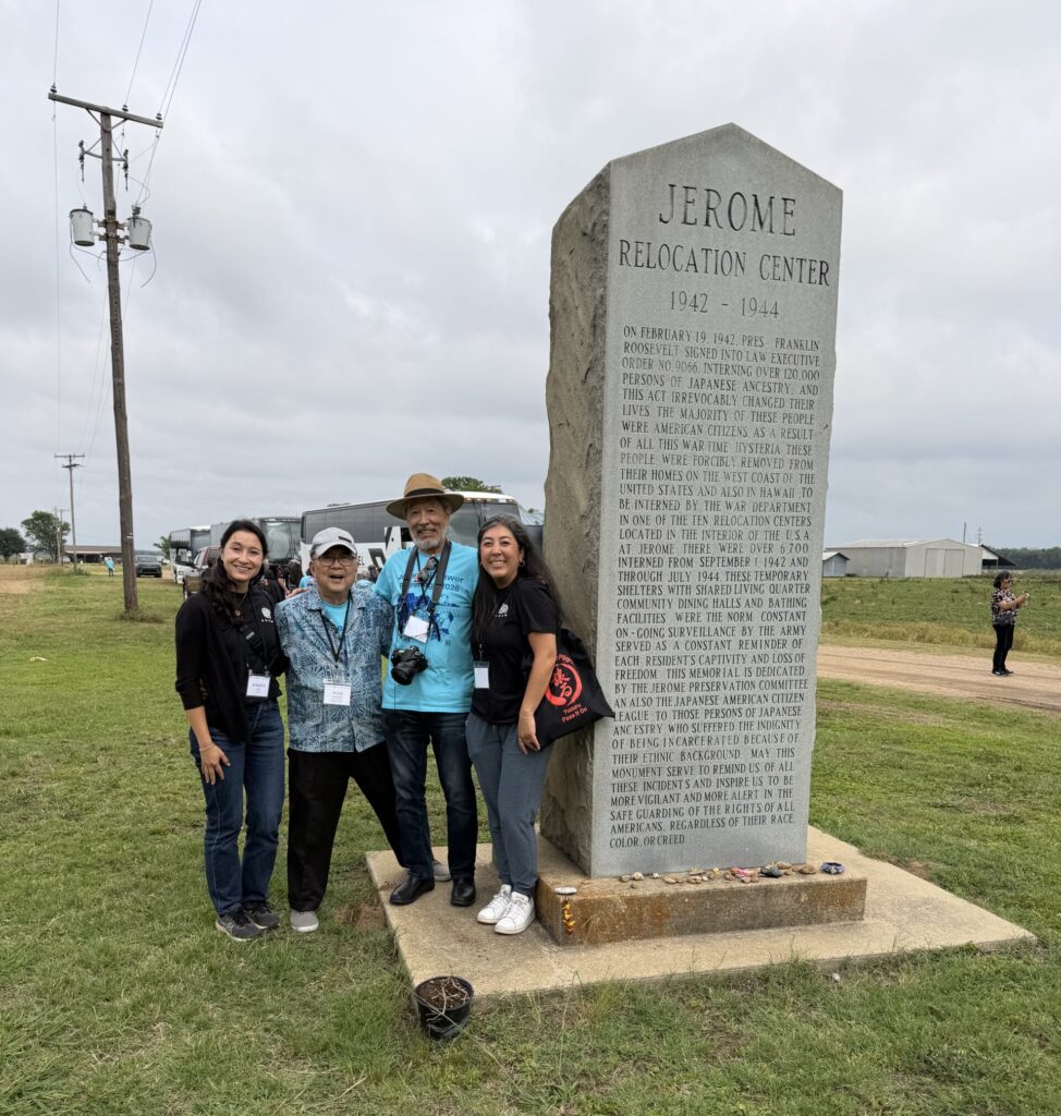 Four people pose smiling in front of the Jerome Relocation Center memorial stone, with tour buses and grain silos visible in the background.