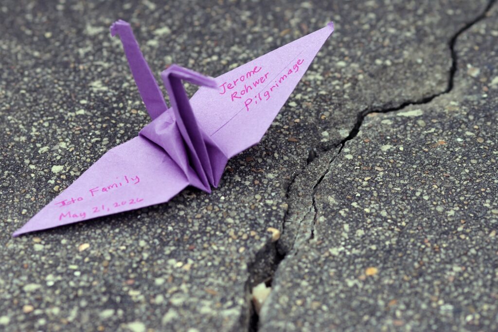 A purple origami paper crane inscribed with "Jerome Rohwer Pilgrimage, Ito Family, May 21, 2026" rests on a cracked paved surface.