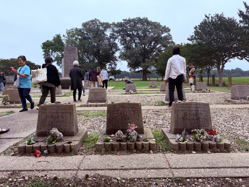 Small children's grave markers, with flowers in front, including three reading "Infant," sit in a cemetery as a group of visitors gathers around a taller memorial in the background.
