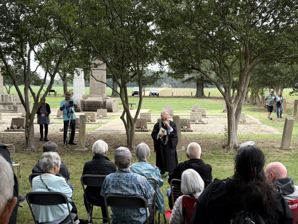 A woman in black robes speaks into a microphone while leading a memorial service among gravestones and trees, with seated attendees facing her.