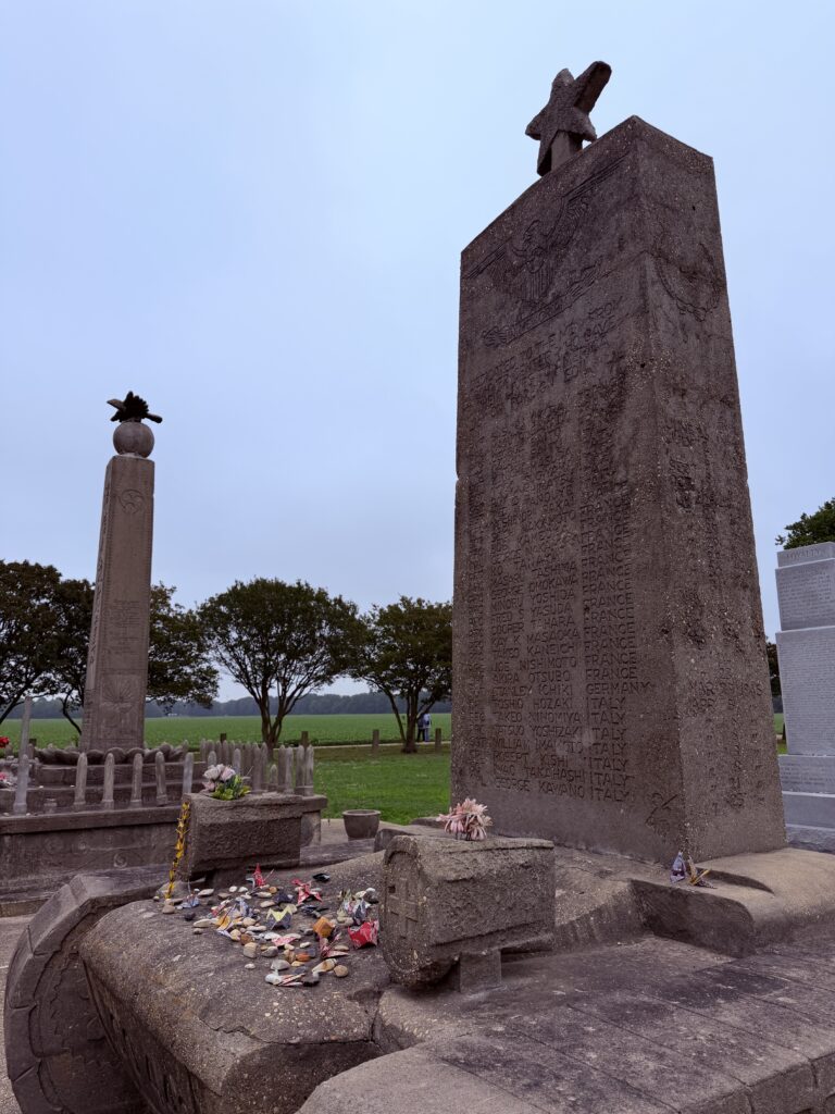 A tall granite memorial engraved with a long list of names honors Japanese American soldiers, standing beside another memorial column decorated with origami cranes and flowers.