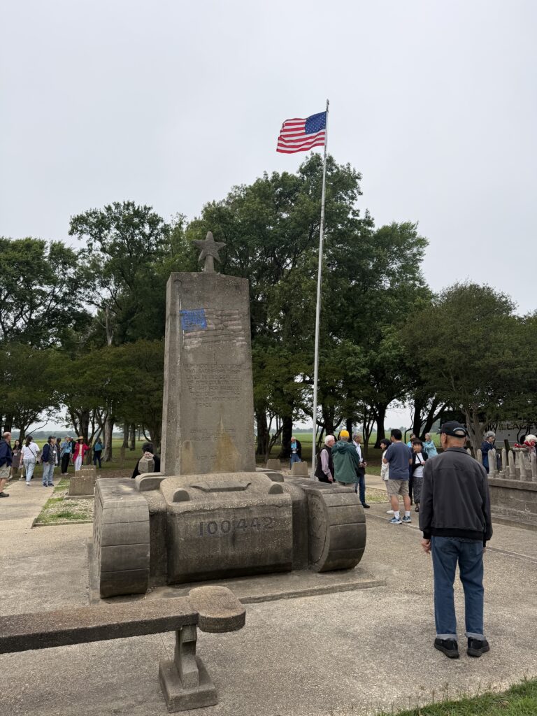 One man in front facing away, and a group of visitors in the background, gather around a stone war memorial topped with a star, beneath a large American flag flying against a cloudy sky.