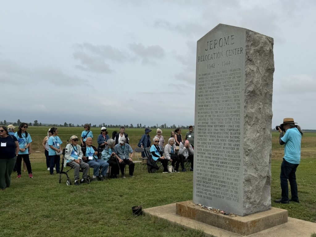 A row of seated elderly attendees, many wearing matching light blue shirts, faces the Jerome Relocation Center memorial stone as a photographer takes their picture in an open field.
