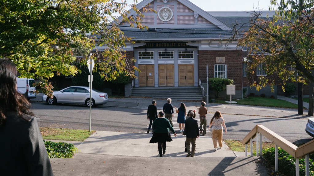 Group of people walking away from camera, crossing a road toward a Japanese temple. Fall trees and blue sky frame the photo.