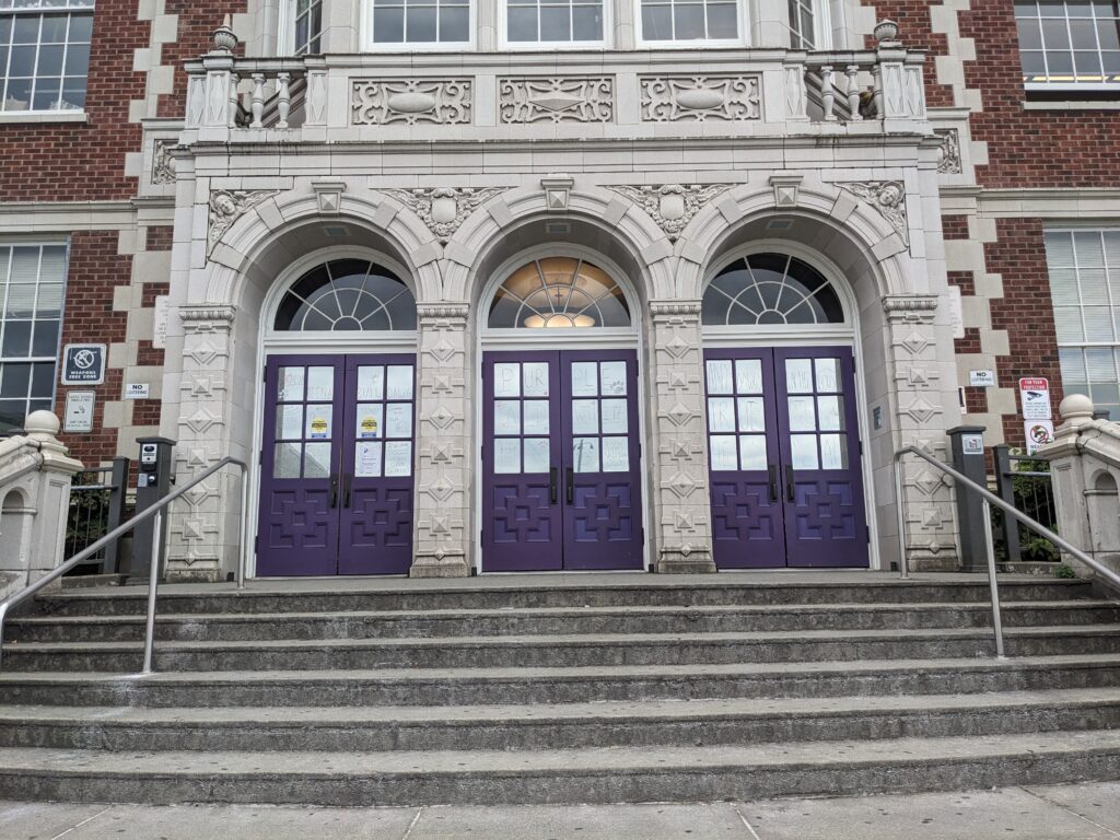 Photo showing the outside front doors of a school. The doors are dark purple, and there are steps leading up to a landing