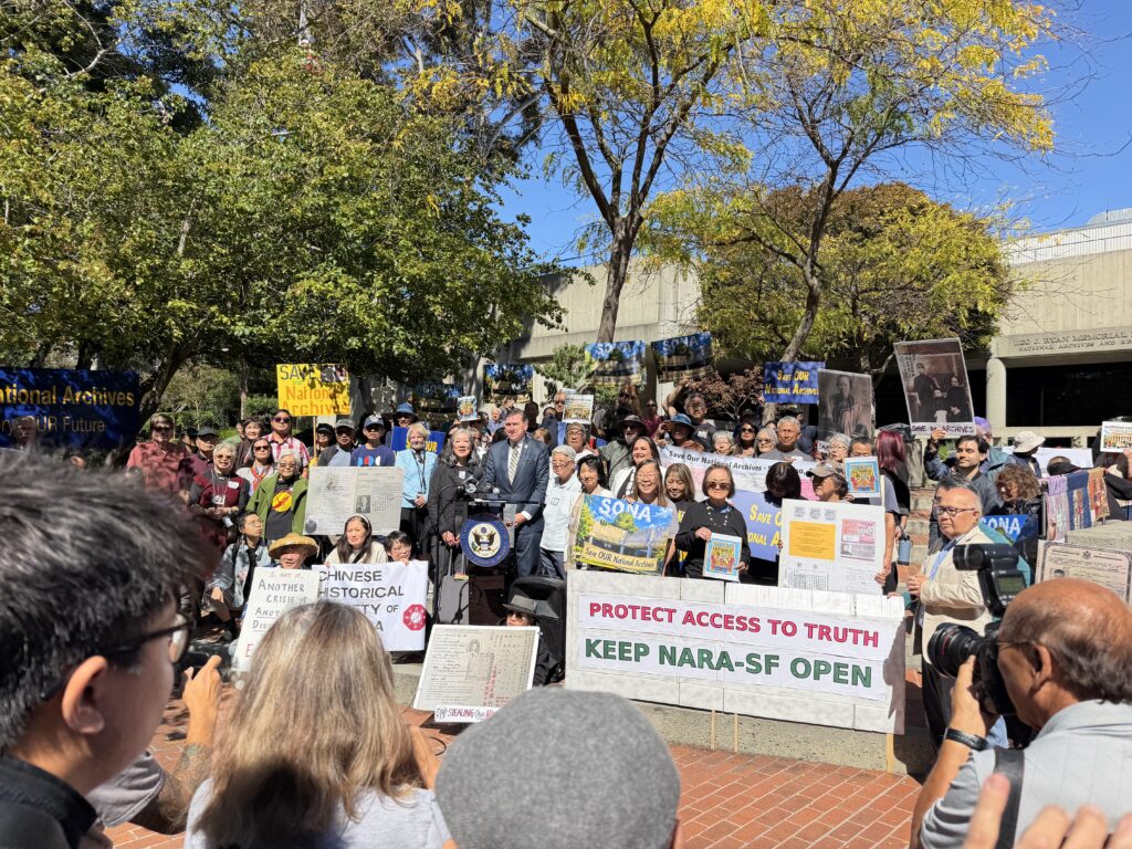 Photo of a large group of people holding protest signs attending a press conference on a sunny day.