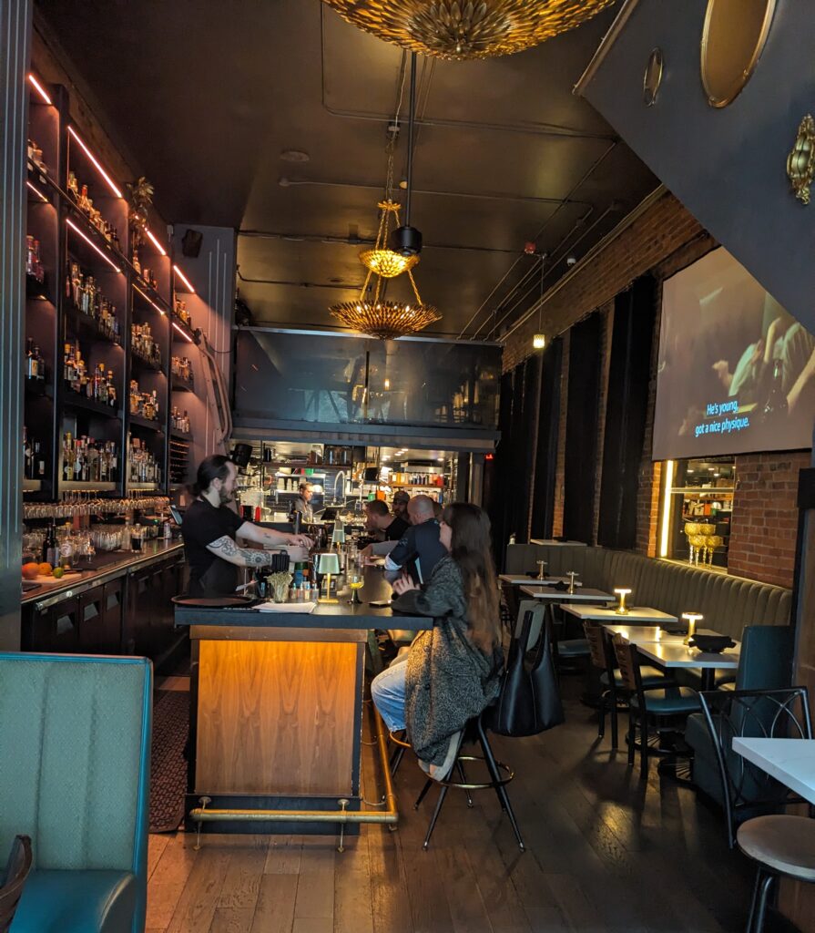 Photo taken inside a bar, showing patrons at the bar and bartender serving drinks