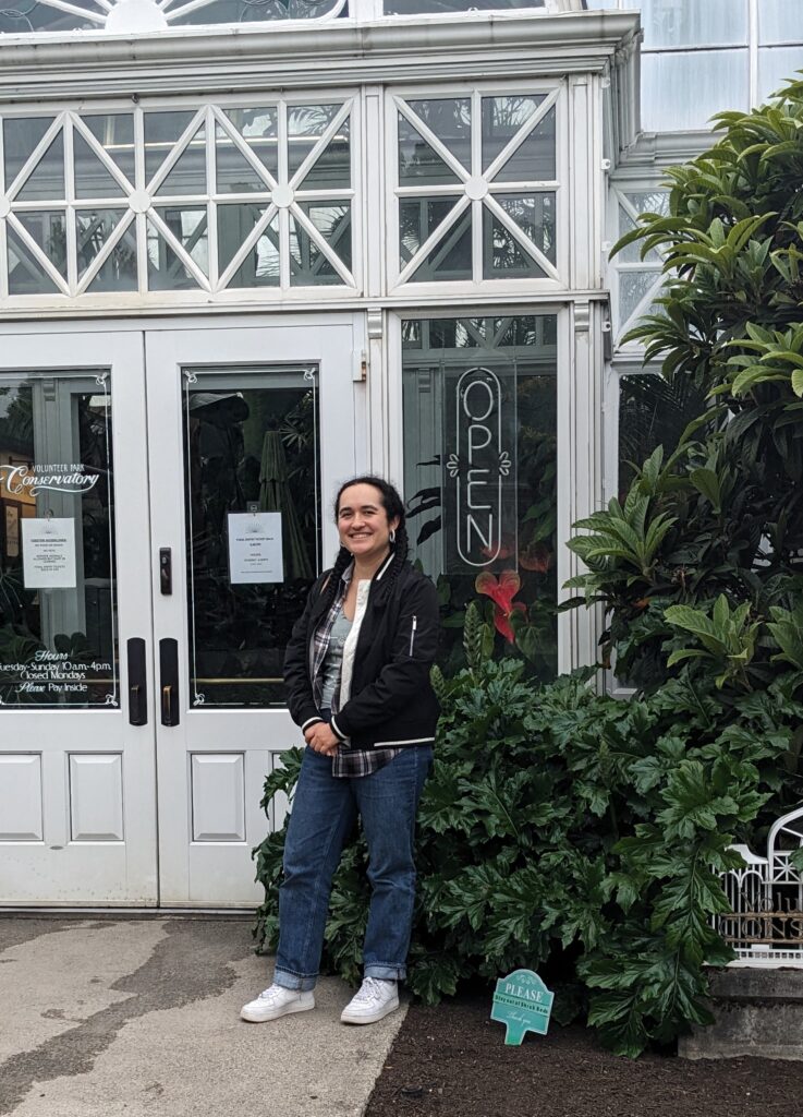 Photo of woman standing outside two white double doors with foliage on her left side