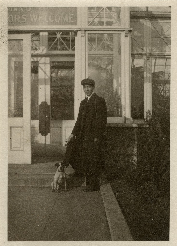 Black and white image of a man standing in front of a conservatory door with a small dog
