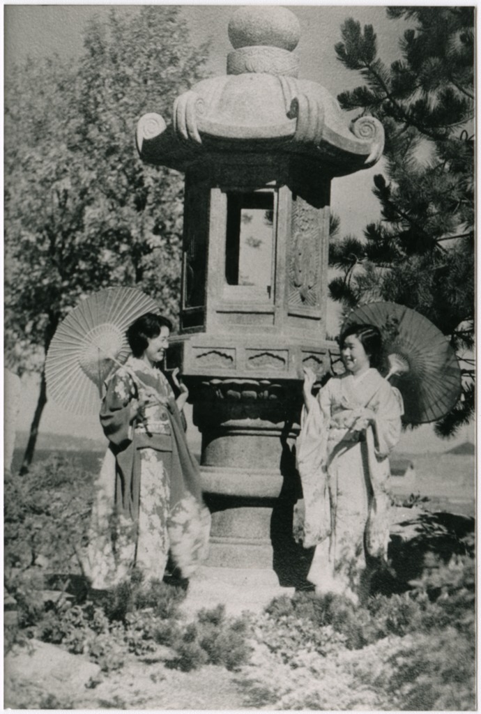 Black and white image of two women dressed in kimono stand in front of a large stone lantern in a park