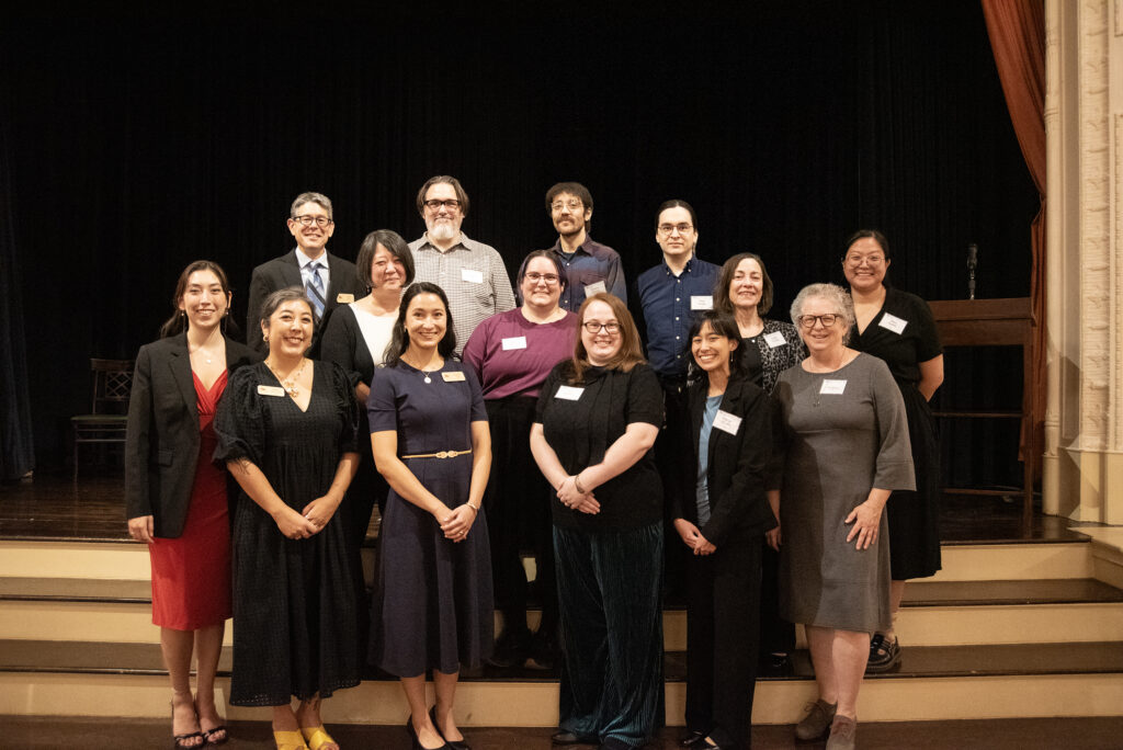 Group photo showing people in business casual attire standing in front of a stage, smiling at the camera